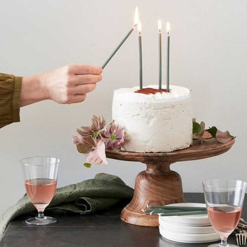 Floral Society Wishing Candles Skinny Birthday Candles being lit on a cake on a wooden cake stand with glasses of wine on the table.